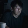 暗い部屋でモニターの光だけに照らされた40代男性。まぶたが垂れ、目の下にクマがあり、口角が下がっている。手前に飲みかけのコーヒー