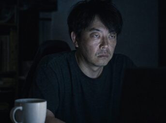 暗い部屋でモニターの光だけに照らされた40代男性。まぶたが垂れ、目の下にクマがあり、口角が下がっている。手前に飲みかけのコーヒー