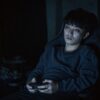暗い部屋で、ゲーミングチェアに深く座りコントローラーを両手で持つ若い男性。画面の光に照らされ、目は充血し下にくまがある