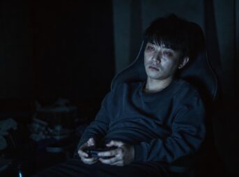 暗い部屋で、ゲーミングチェアに深く座りコントローラーを両手で持つ若い男性。画面の光に照らされ、目は充血し下にくまがある