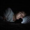 真っ暗な寝室。横になった男性の顔の下半分だけが、手に持ったスマートフォンの光に照らされている