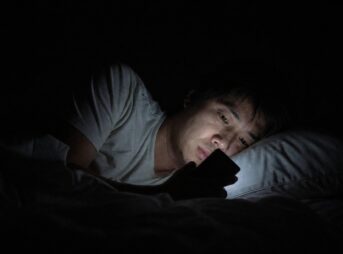 真っ暗な寝室。横になった男性の顔の下半分だけが、手に持ったスマートフォンの光に照らされている