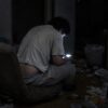 散らかった暗い部屋で床の座椅子にもたれて座り、スマートフォンをのぞき込む若い男性のうしろ姿。ずり下がったズボンから下着がのぞき、周りに菓子の袋やティッシュが散乱している