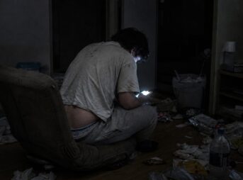 散らかった暗い部屋で床の座椅子にもたれて座り、スマートフォンをのぞき込む若い男性のうしろ姿。ずり下がったズボンから下着がのぞき、周りに菓子の袋やティッシュが散乱している